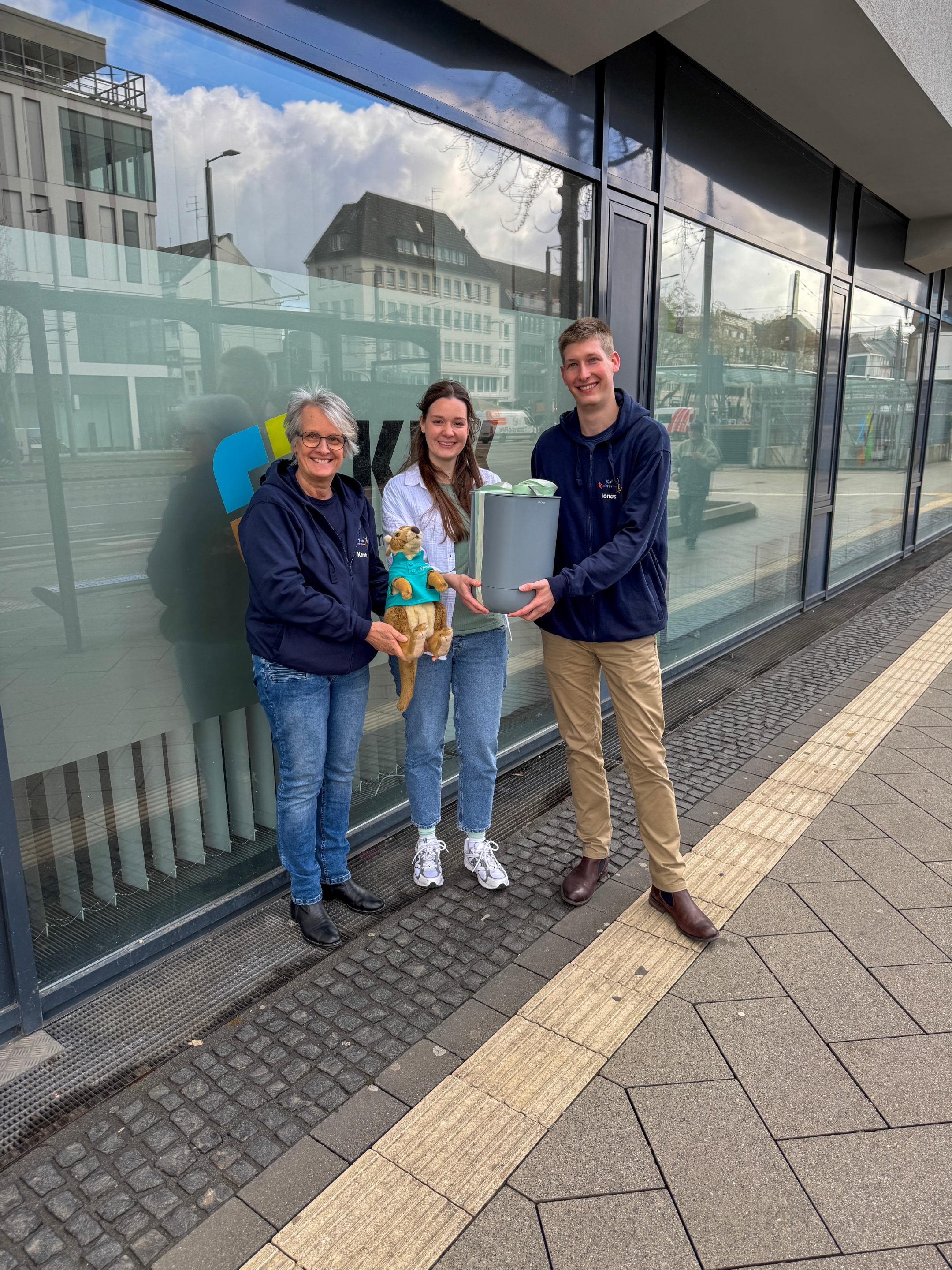 Vor einer Fensterfront auf dem Bürgersteig sind drei Personen, zwei Frauen und ein Mann. (Von rechts nach links) Eine Frau mit einem dunkelblauen Hoodie, eine Frau mit hellblauem geöffneten Hemd, ein Mann mit einem dunkelblauen Hoodie. Die Frau in der Mitte überreicht dem Mann einen grauen Kanister, der Bokashi. Kängu ist auch auf dem Foto in der Hand der Frau mit dem dunklen Hoodie.