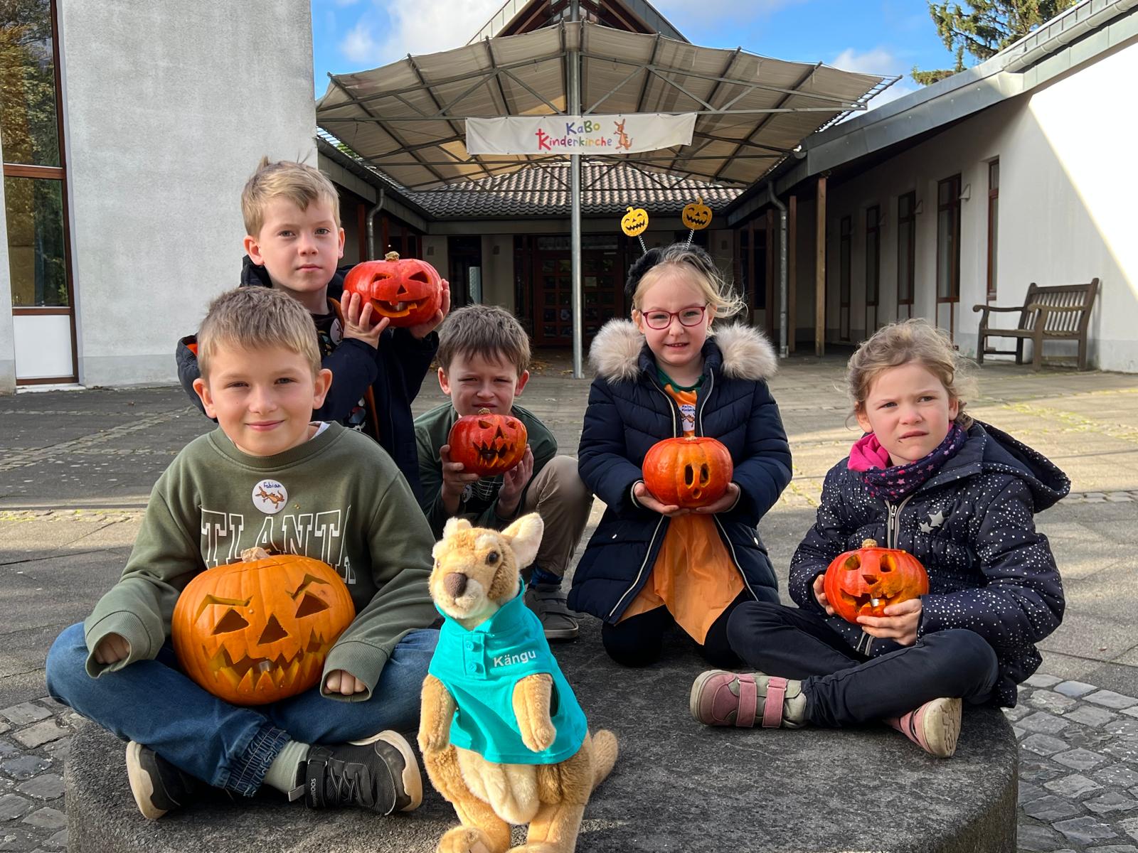 Viele Kinder mit je einem Kürbis und Kängu sitzen auf einem Stein. Im Hintergrund das Pfarrzentrum von St. Karl Borromäus.