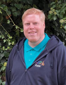 Ein Mann, der lacht,  ist im Vordergrund. Er hat eine dunkelblaue Jacke an und trägt ein Tshirt in der Farbe türtkis. Er hat blonde Haare. Im Hintergrund ist eine dunkelgrüne Hecke. Es ist nur der Teil bis Mitte des Körpers erkennbar.