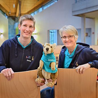 Zwei Personen sitzen auf Stühlen und schauen in die Kamera. Ein Mann (links) und eine Frau (rechts). In der Mitte halten beide das Känguru, Kängu fest.