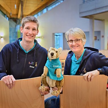Zwei Personen sitzen auf Stühlen und schauen in die Kamera. Ein Mann (links) und eine Frau (rechts). In der Mitte halten beide das Känguru, Kängu fest.