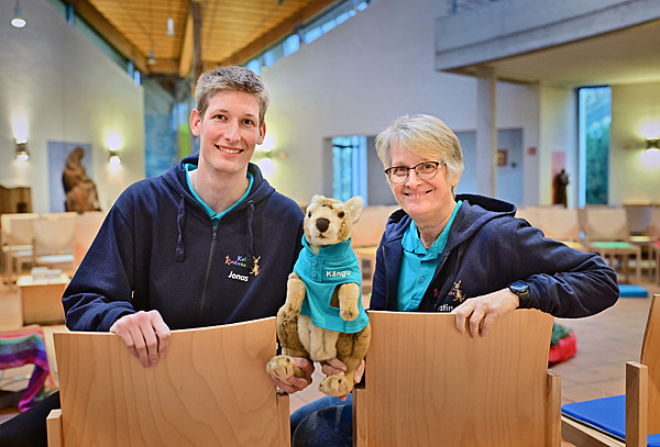 Zwei Personen sitzen auf Stühlen und schauen in die Kamera. Ein Mann (links) und eine Frau (rechts). In der Mitte halten beide das Känguru, Kängu fest.