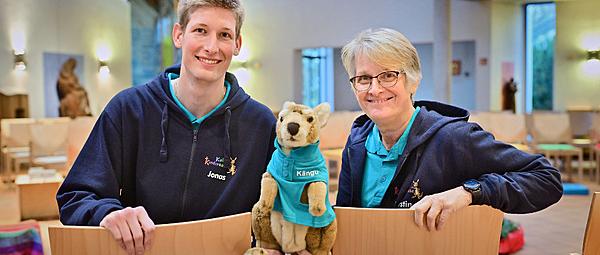 Zwei Personen sitzen auf Stühlen und schauen in die Kamera. Ein Mann (links) und eine Frau (rechts). In der Mitte halten beide das Känguru, Kängu fest.