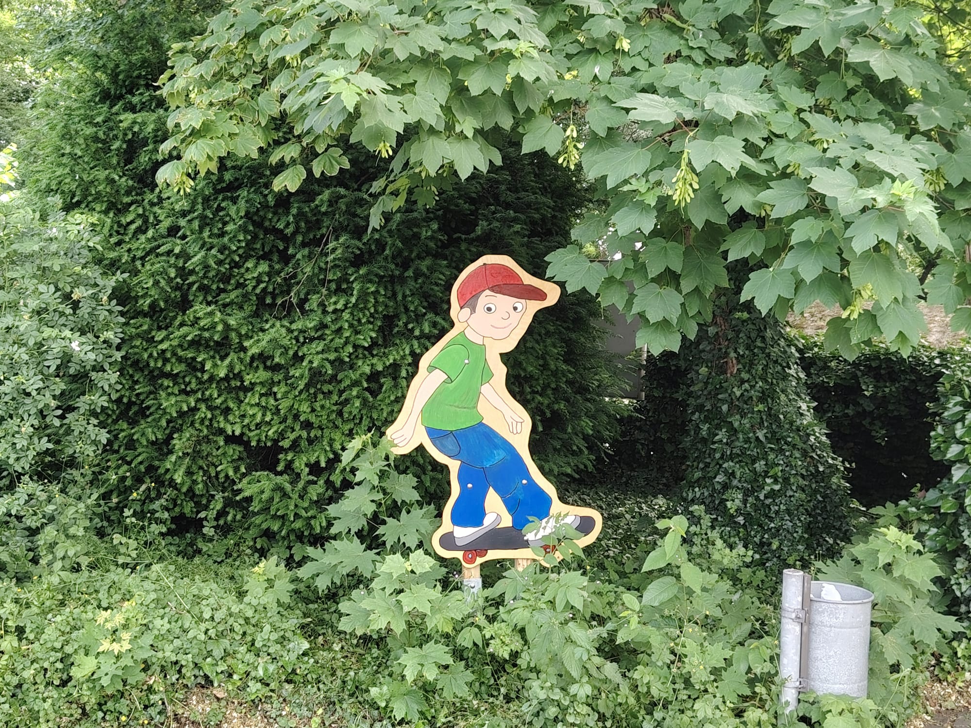 Im Grünen ist ein Junge auf einem Skateboard mit einer roten Käppi, grünem T-Shirt und einer blauen Hose.