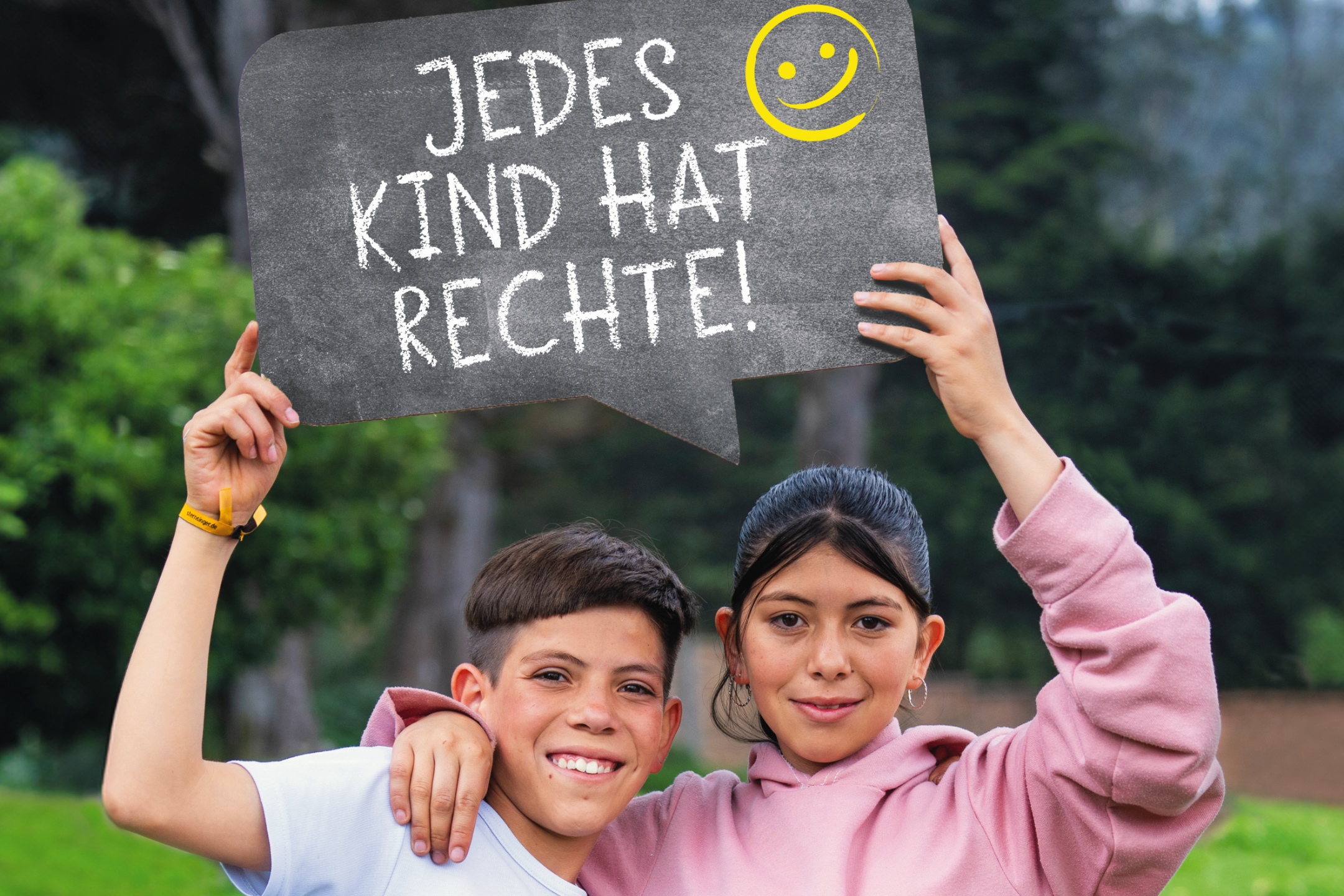 Zwei Kinder (ein Junge und ein Mädchen) halten ein Schild mit der Aufschrift 'Jedes Kind hat Rechte!' hoch. Der Junge trägt ein weißes T-shirt, das Mädchen ein rosa Pulli. Im Hintergrund sind ein Bau und eine grüne Hecke sowie eine hellgrüne Wiese.