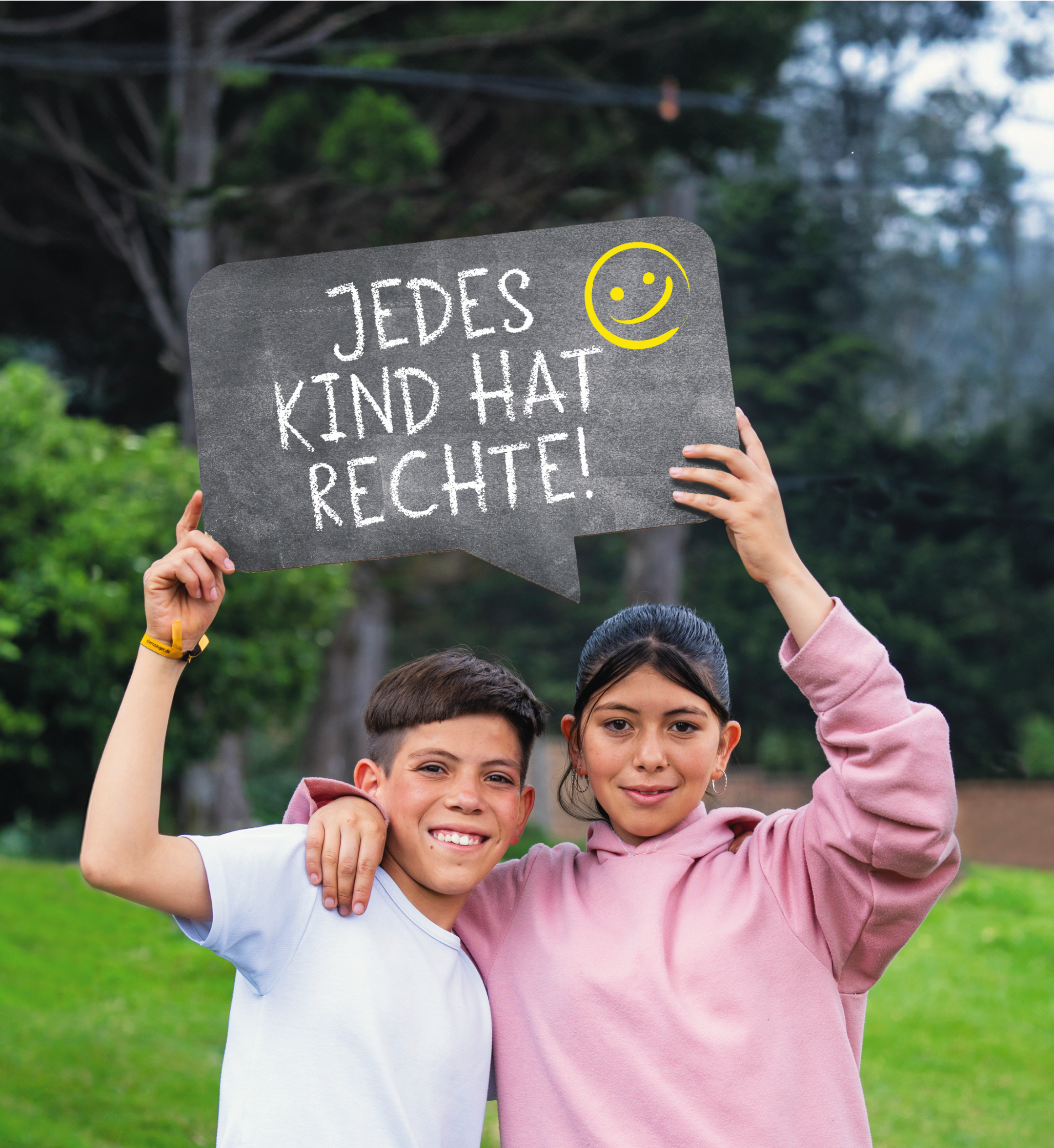 Zwei Kinder (ein Junge und ein Mädchen) halten ein Schild mit der Aufschrift 'Jedes Kind hat Rechte!' hoch. Der Junge trägt ein weißes T-shirt, das Mädchen ein rosa Pulli. Im Hintergrund sind ein Bau und eine grüne Hecke sowie eine hellgrüne Wiese.