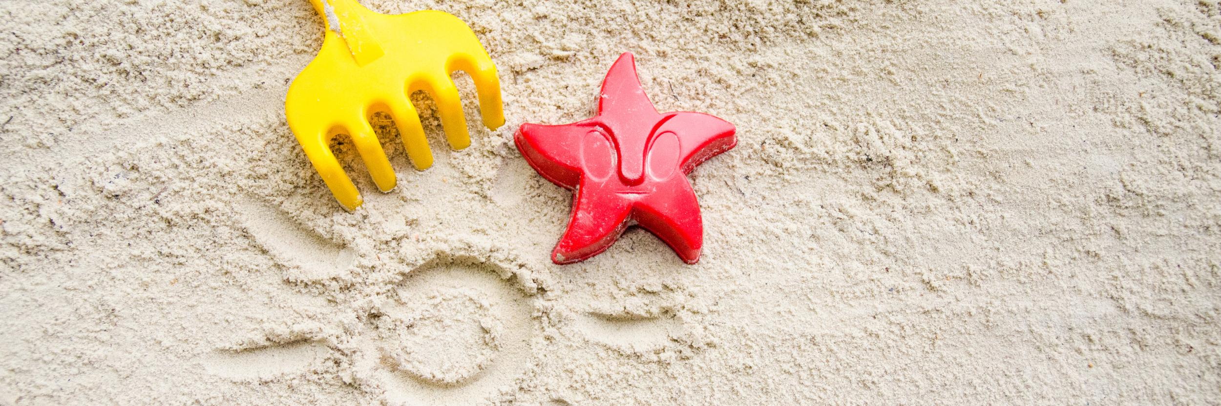 Ein gelbes Rechen und ein rotes Austechförmchen in Form eines Seesterns auf Sand. In den Sand ist eine Sonne mit Strahlen gemalt