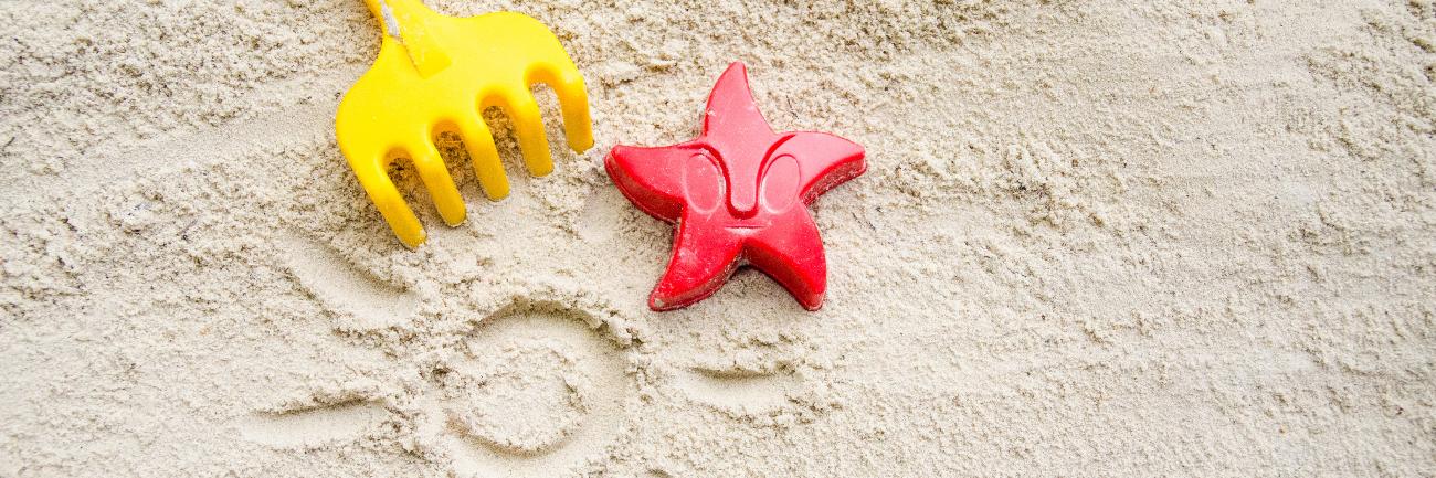 Ein gelbes Rechen und ein rotes Austechförmchen in Form eines Seesterns auf Sand. In den Sand ist eine Sonne mit Strahlen gemalt