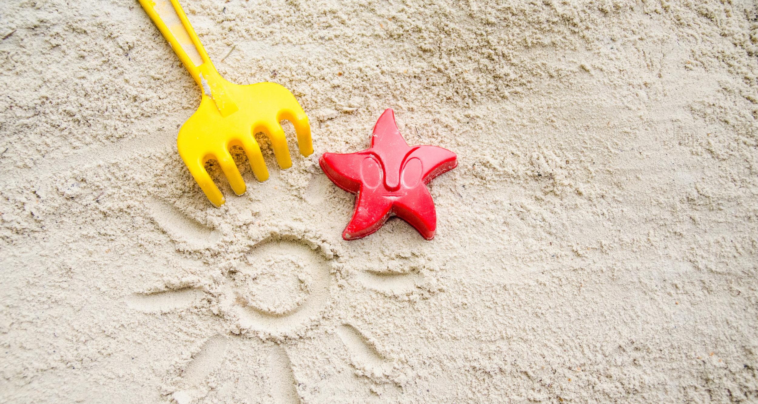Ein gelbes Rechen und ein rotes Austechförmchen in Form eines Seesterns auf Sand. In den Sand ist eine Sonne mit Strahlen gemalt