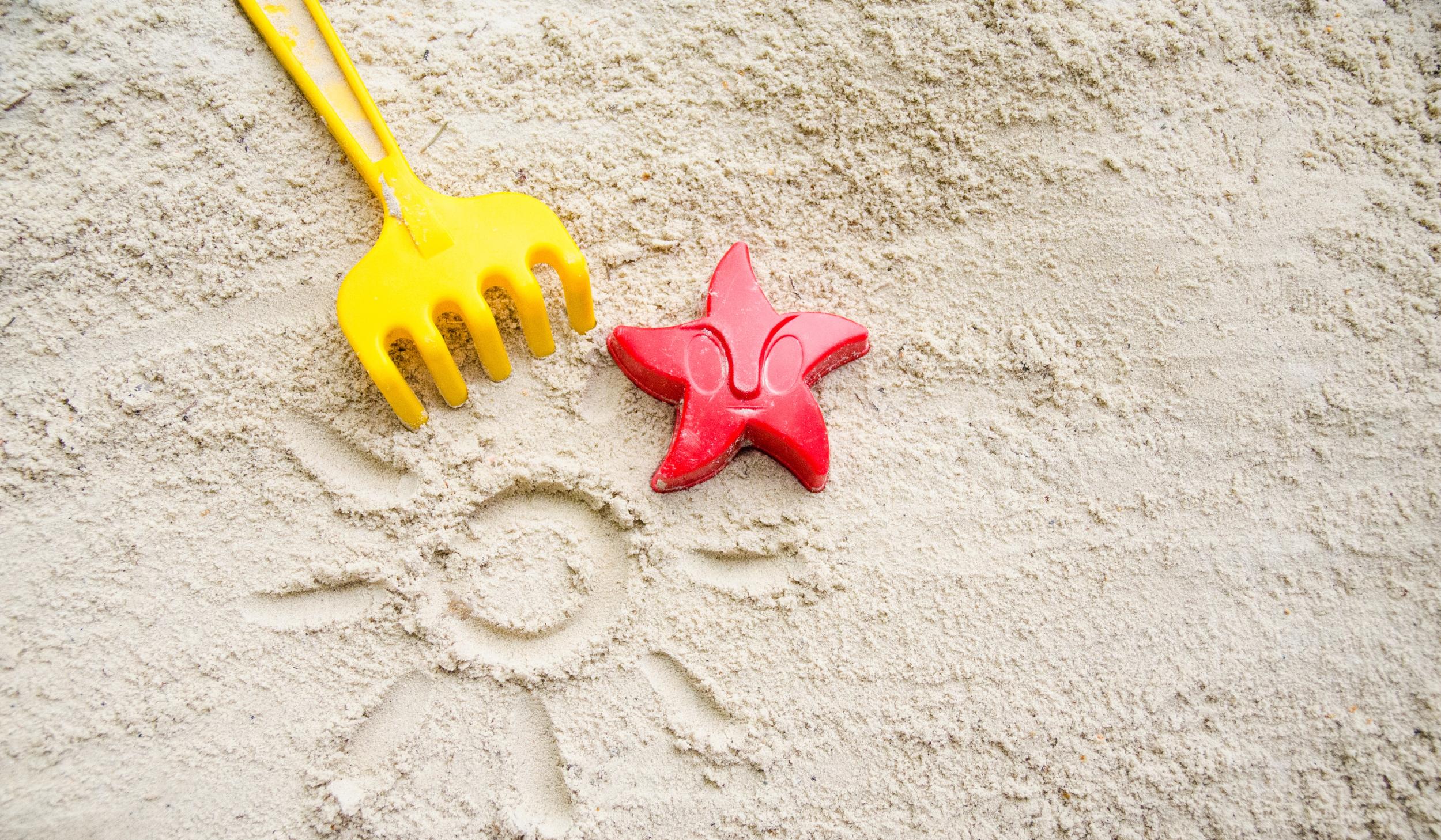Ein gelbes Rechen und ein rotes Austechförmchen in Form eines Seesterns auf Sand. In den Sand ist eine Sonne mit Strahlen gemalt