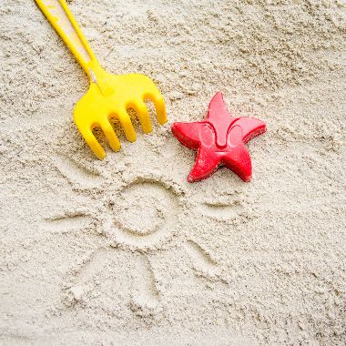 Ein gelbes Rechen und ein rotes Austechförmchen in Form eines Seesterns auf Sand. In den Sand ist eine Sonne mit Strahlen gemalt
