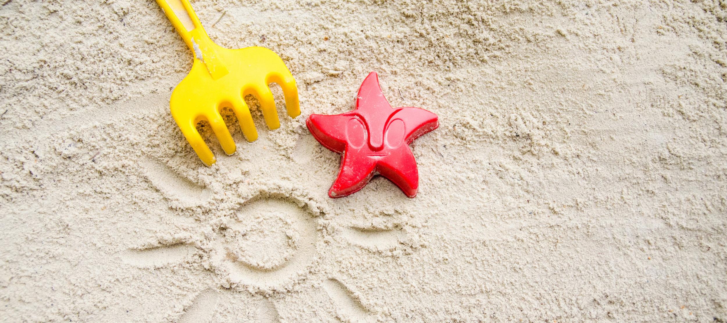 Ein gelbes Rechen und ein rotes Austechförmchen in Form eines Seesterns auf Sand. In den Sand ist eine Sonne mit Strahlen gemalt