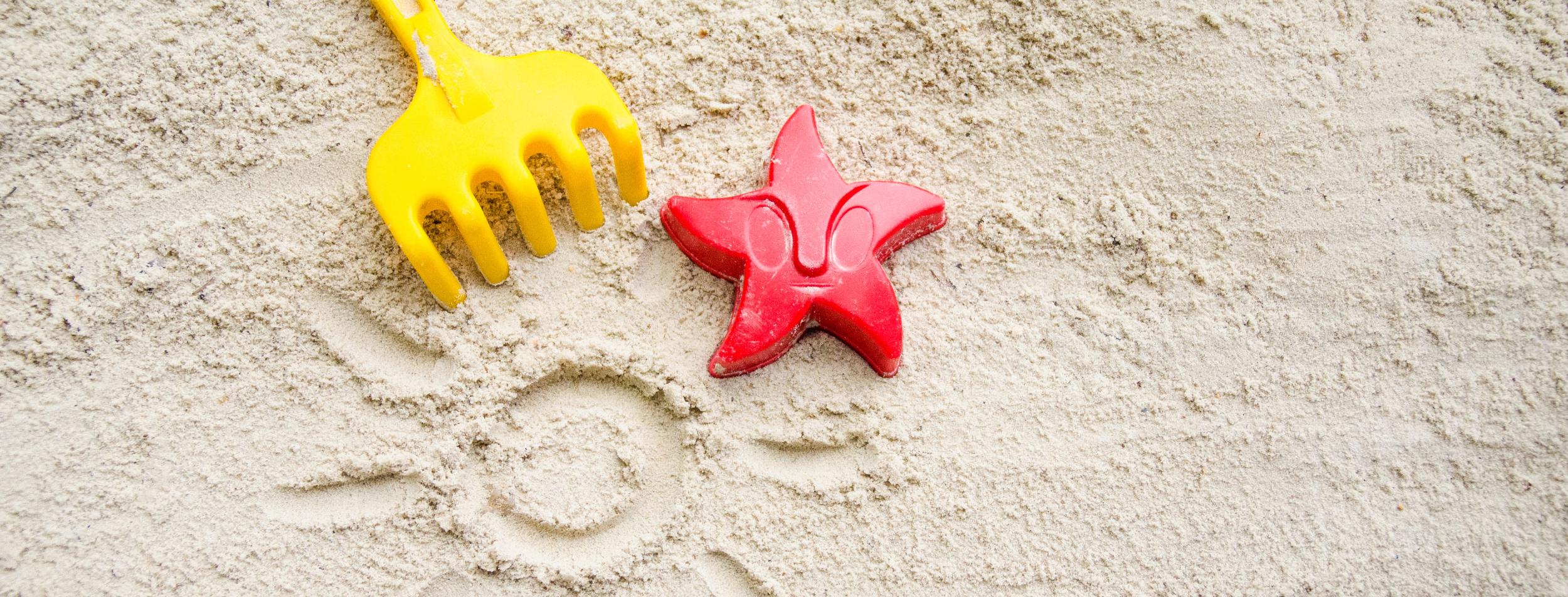 Ein gelbes Rechen und ein rotes Austechförmchen in Form eines Seesterns auf Sand. In den Sand ist eine Sonne mit Strahlen gemalt