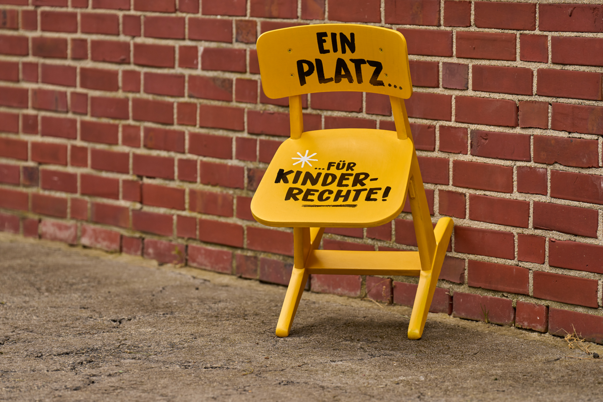 Ein gelber Stuhl lehnt an einer Hauswand. Auf dem Stuhl findet sich die Aufschrift 'Ein Platz...' und 'für Kinderrechte'.