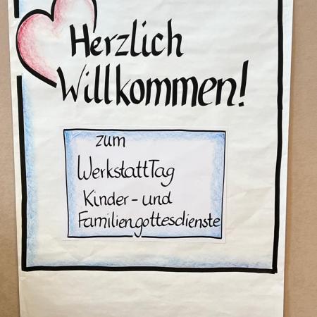 Auf einem hellbraunen Hintergrund hängt ein weißes Papierflipchart, auf diesem in einem schwarzen Rahmen gezeichnet ein rotes Herz und Herzlich Willkommen sowie ein blauer Kasten mit 'zu WerkstattTag Kinder- und Familiengottesdienste'.