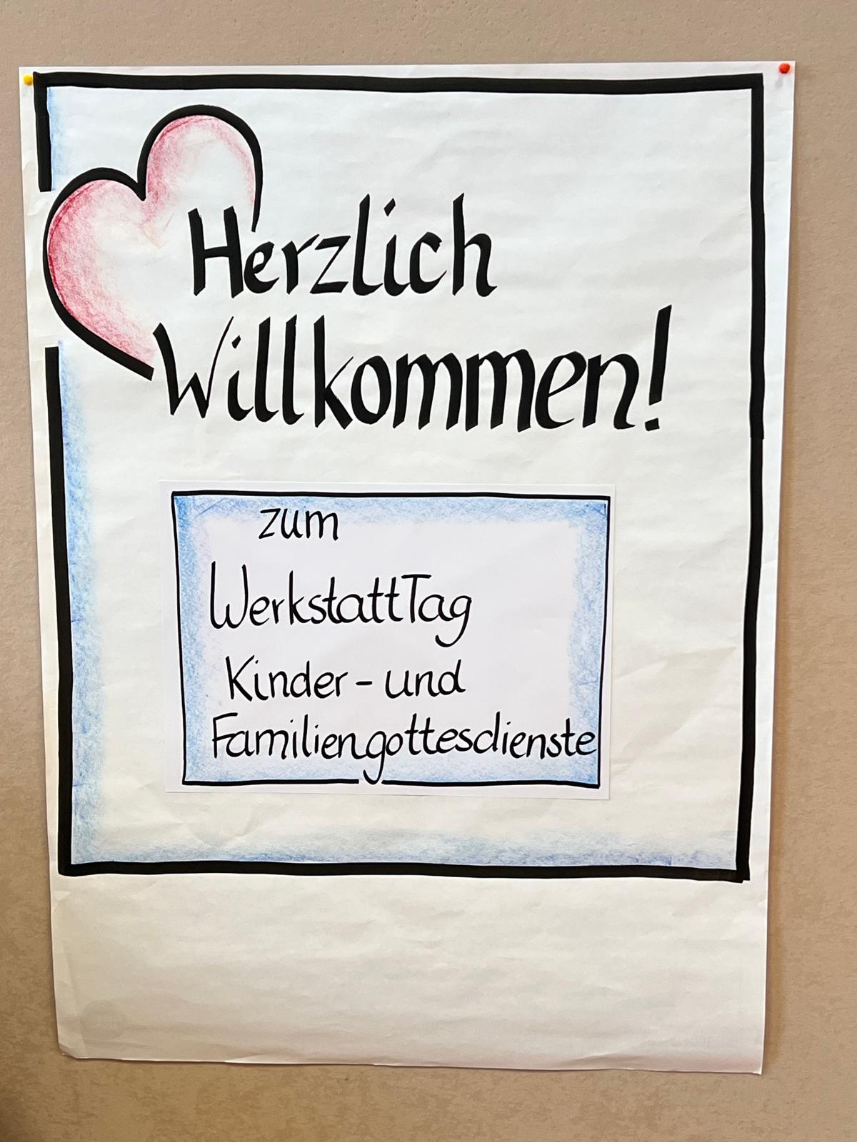 Auf einem hellbraunen Hintergrund hängt ein weißes Papierflipchart, auf diesem in einem schwarzen Rahmen gezeichnet ein rotes Herz und Herzlich Willkommen sowie ein blauer Kasten mit 'zu WerkstattTag Kinder- und Familiengottesdienste'.